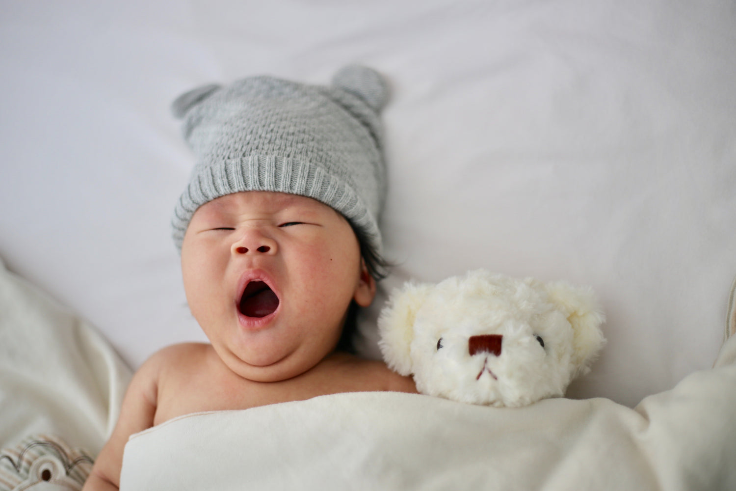Un bébé qui baille allongé sur un lit avec un ourson en peluches | Babykare.fr
